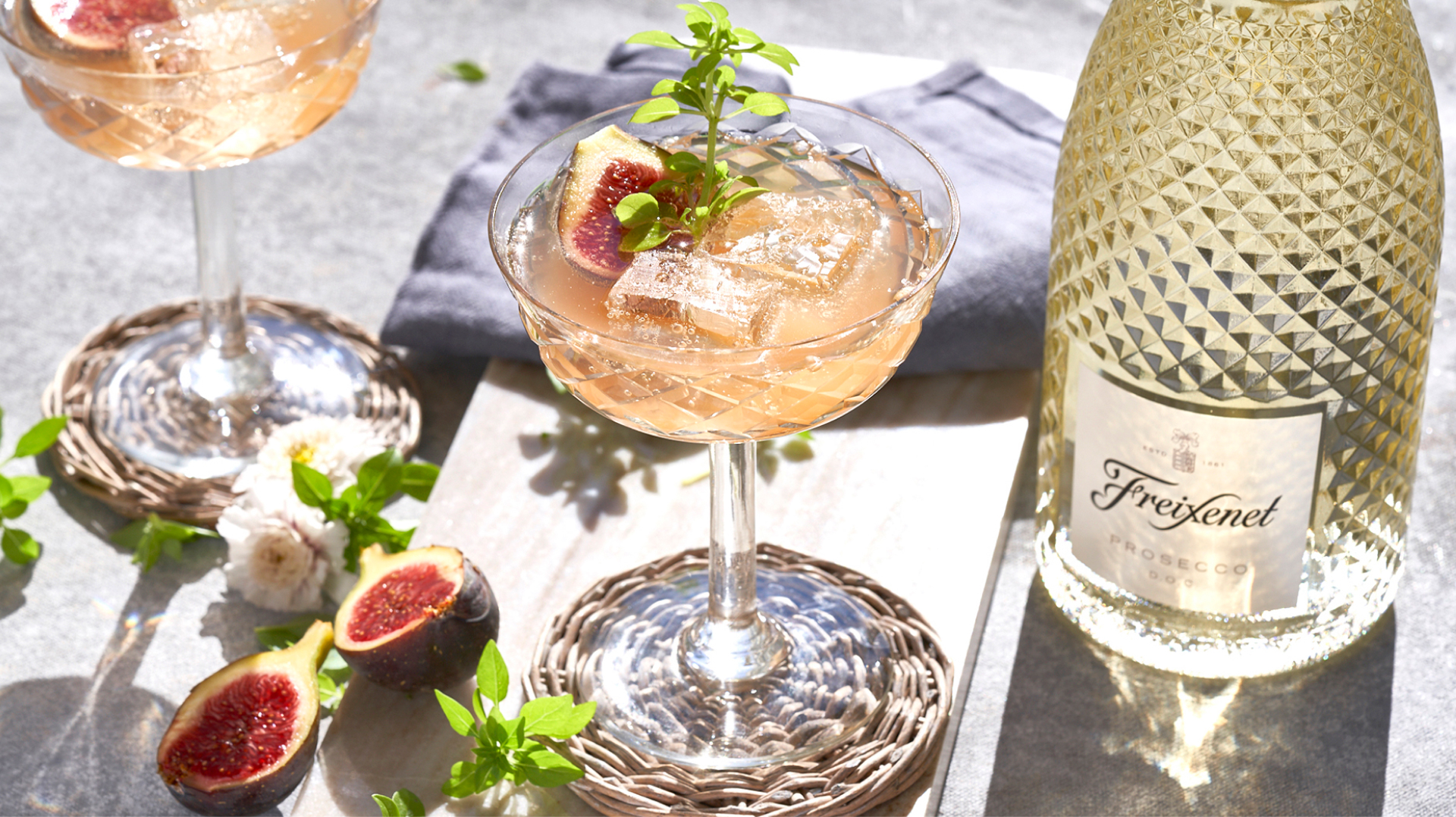 Luminous faceted prosecco bottle alongside two filled coupe glasses with figs and leaves as garnish. Link to recipes and cocktails.