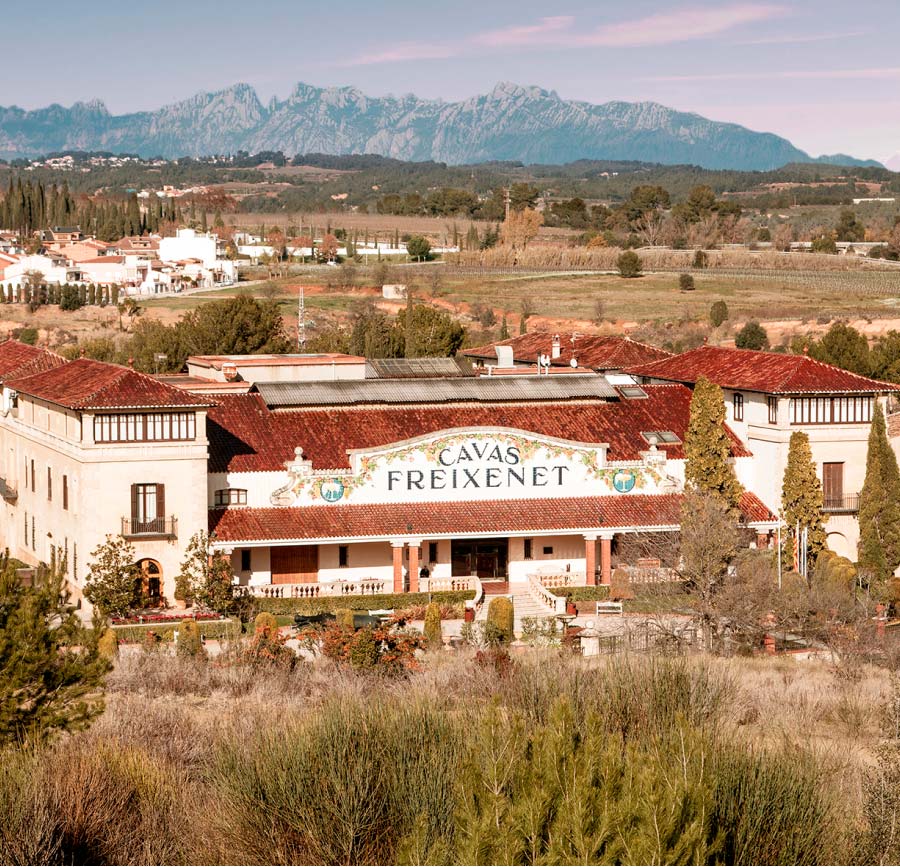 Freixenet Cava headquarters, a large white building with a red roof in the distance. Behind are vineyards, farmland and mountains.