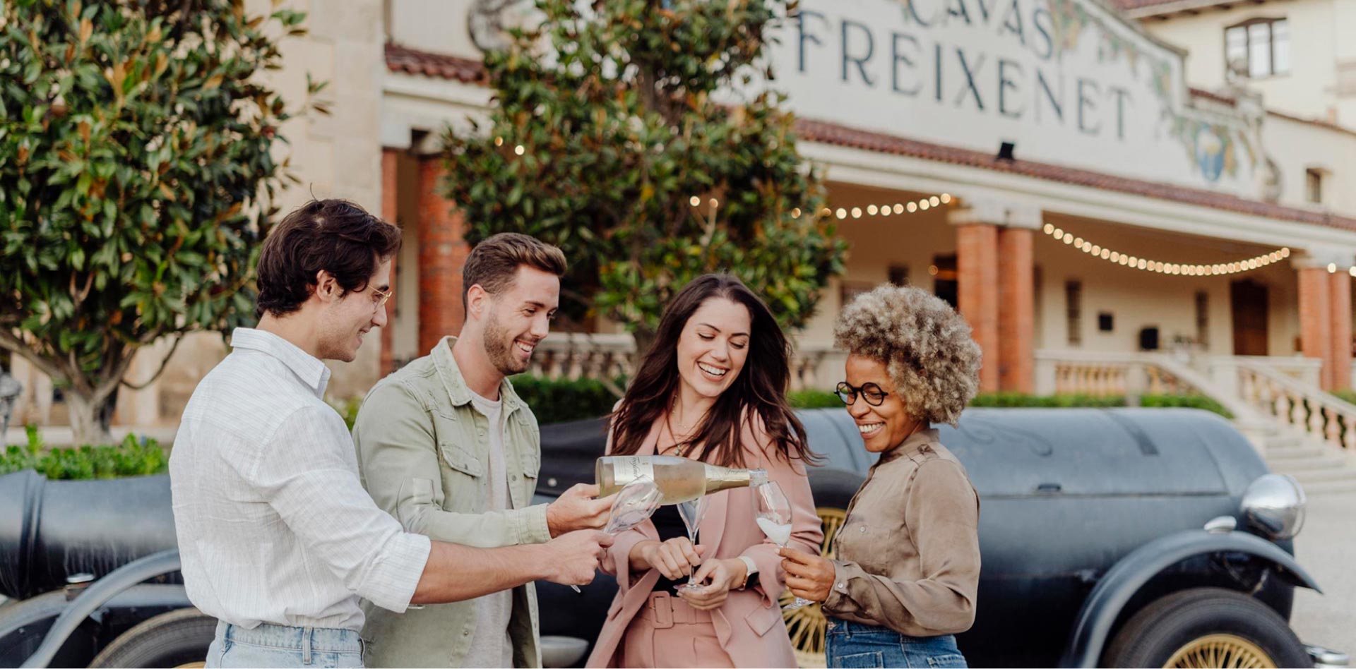 A group of friends, two men and two women, stand beside the bottle-car serving Freixenet into glasses.