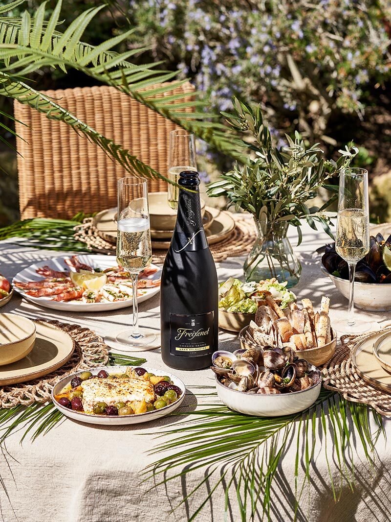 Table set with Cava bottle in the center. Iberian ham and cheeses on plates with abundant foliage nearby.