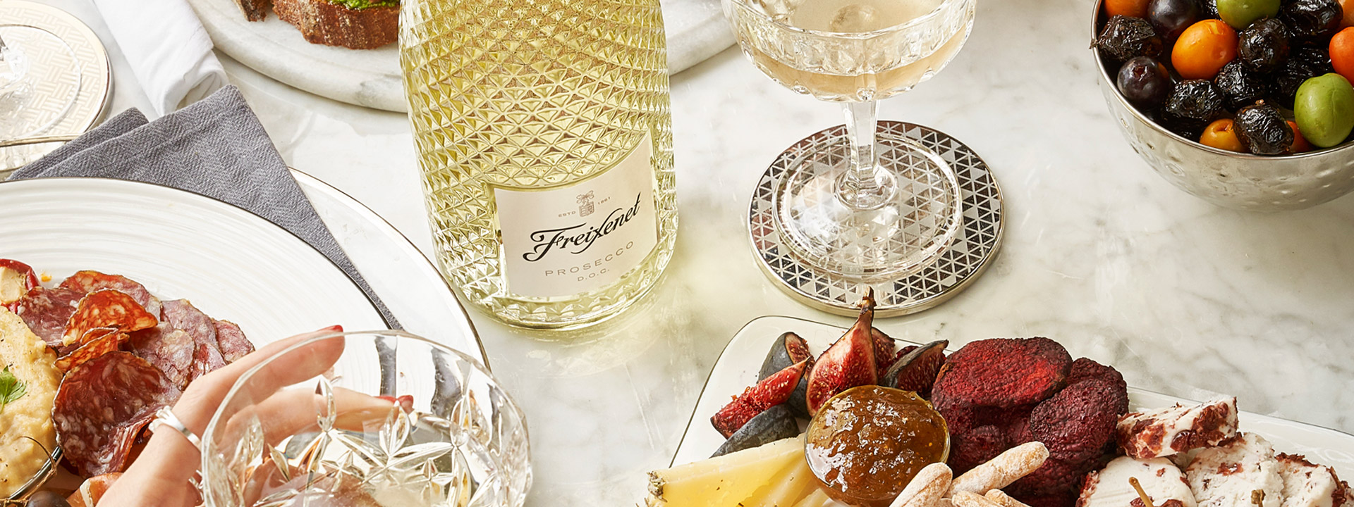 Two women's hands, charcuterie, a faceted sparkling wine bottle and filled coupe glasses.