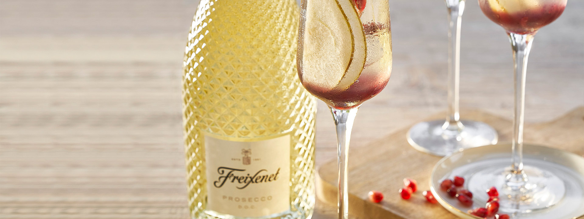 Served cocktails in straight-sided glasses garnished with fruit peels and pomegranite seeds with the faceted Prosecco bottle behind.