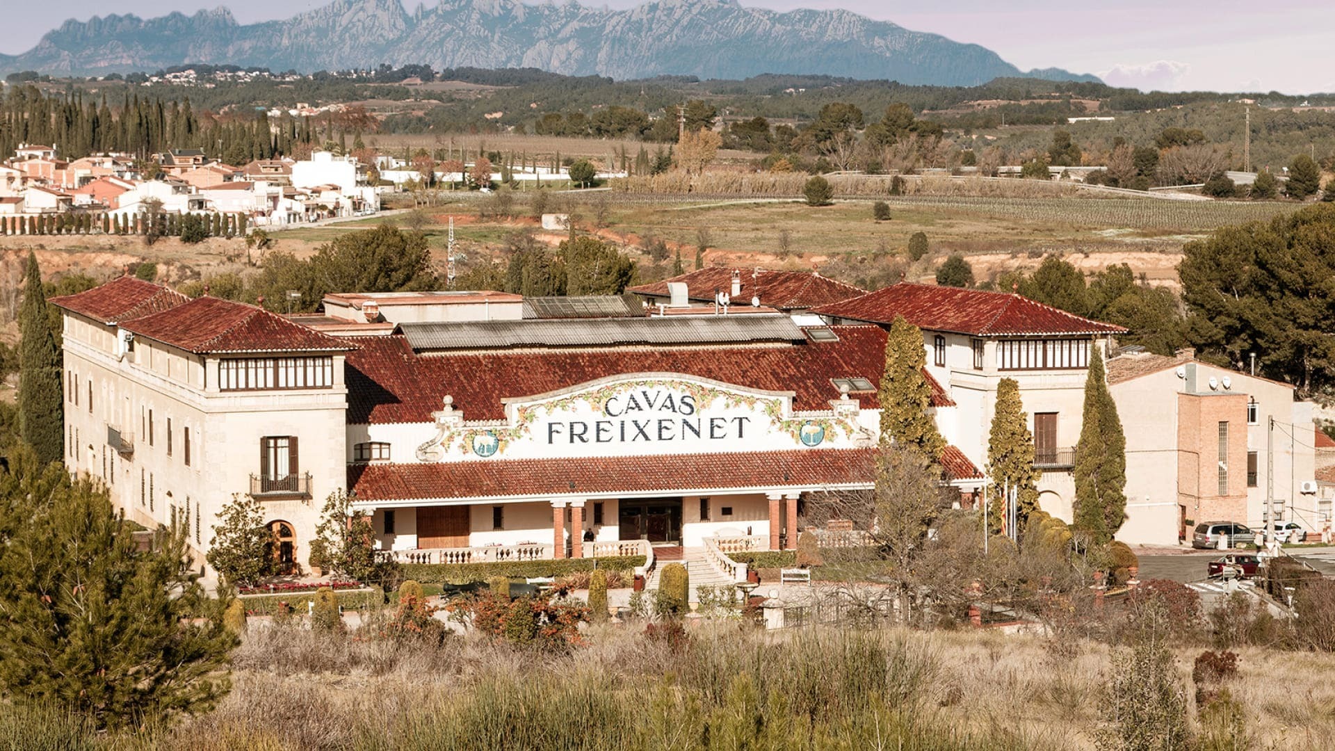 Freixenet Cava headquarters, a large white building with a red roof in the distance. Behind are vineyards, farmland and mountains.