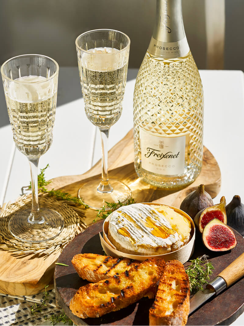 Prosecco bottle with served glasses as if for an outdoor toast, with cheese, bread and figs.