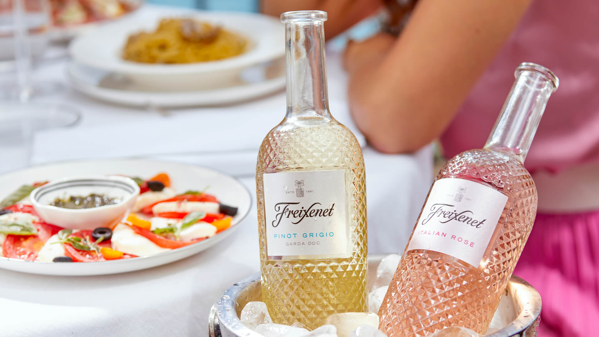 Faceted bottles of white and rosé Freixenet in an ice bucket with a caprese salad behind. Link opens to online store.