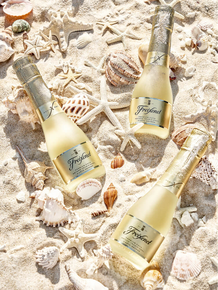 Three individual bottles of Freixenet posed on the sand with shells and starfish in between.