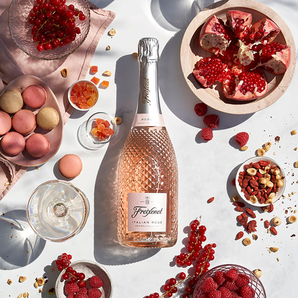 A table with a faceted bottle of Italian Rosé among other pink food items such as a pomegranate, candied fruit, dried berries and macarons shot from above.