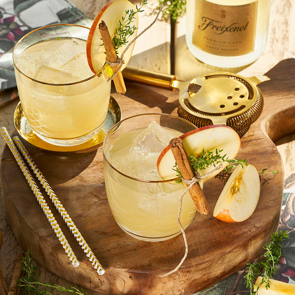 Two served cocktails in tumblers on a wooden board garnished with apple, cinnamon and herbs.