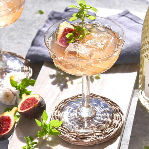A coupe glass with a pink sparkling cocktail garnished with figs and herbs.