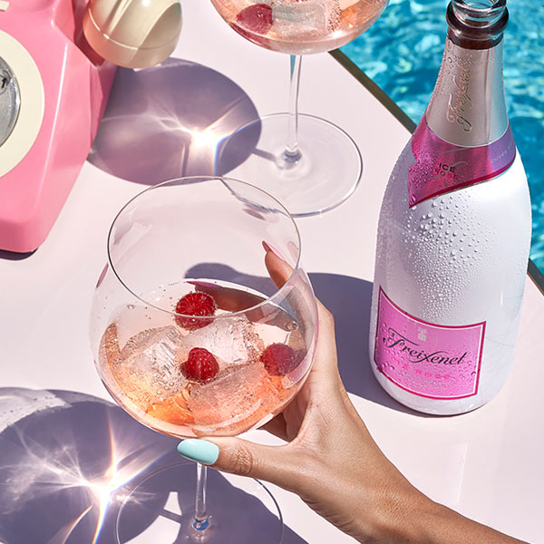 A woman’s hand holding a glass of pink liquid garnished with berries, poolside. The bottle is also visible. 