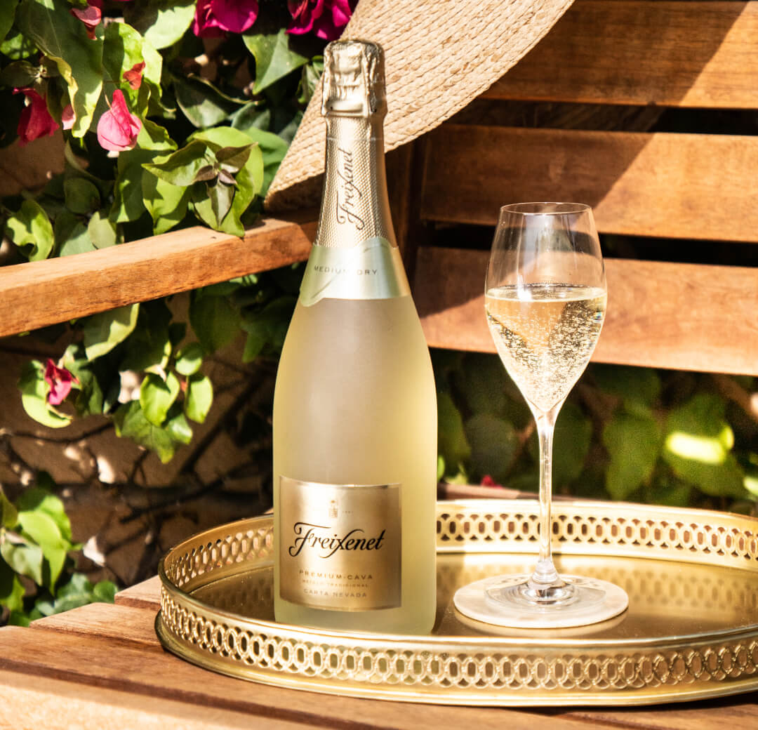 Bottle of Freixenet Cava beside a served glass on a metal tray, on a wooden bench near a blooming bush.