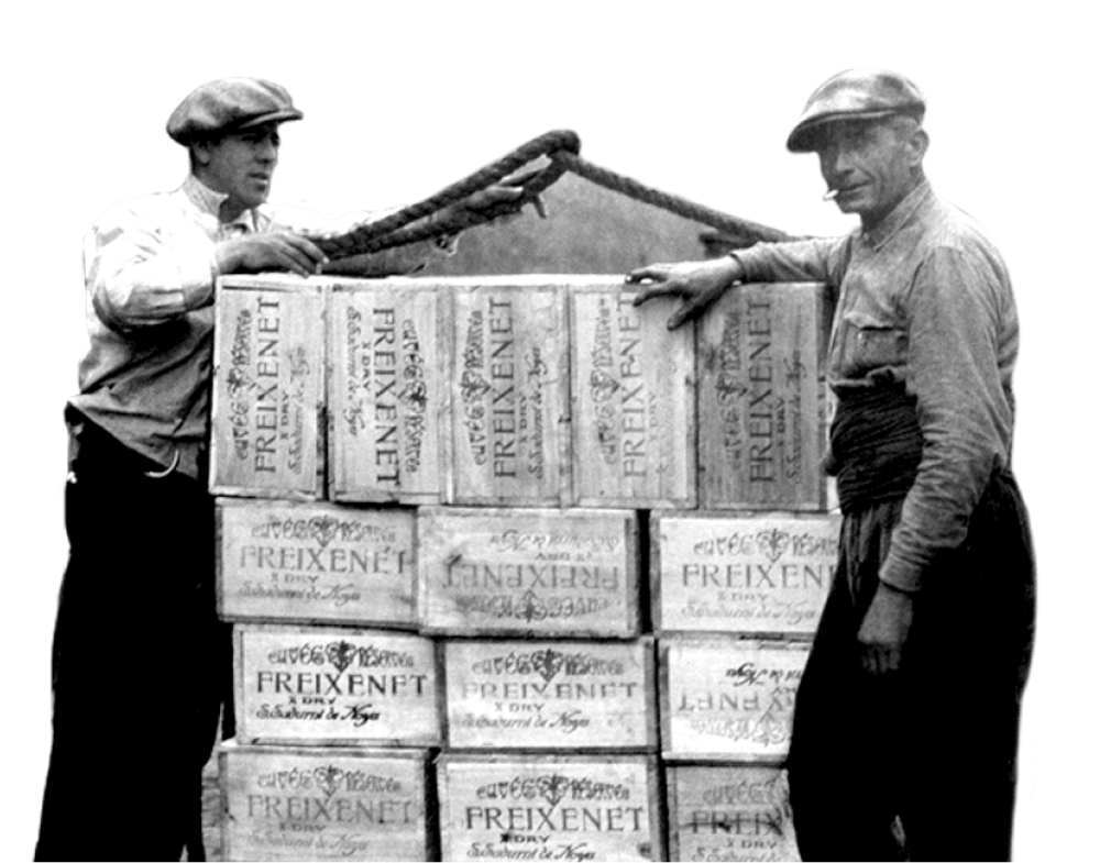 Two men with flat caps pose in front of crates of Freixenet.