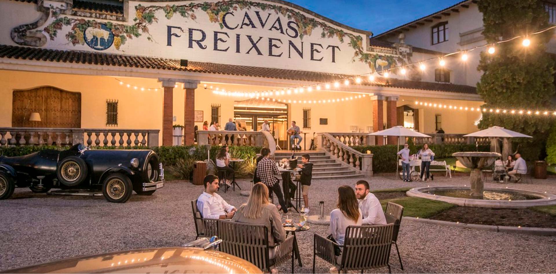 Freixenet Cava headquarters at night with a fountain in front and strung lights in the evening. Groups of people congregate at tables and walk up the stairs.