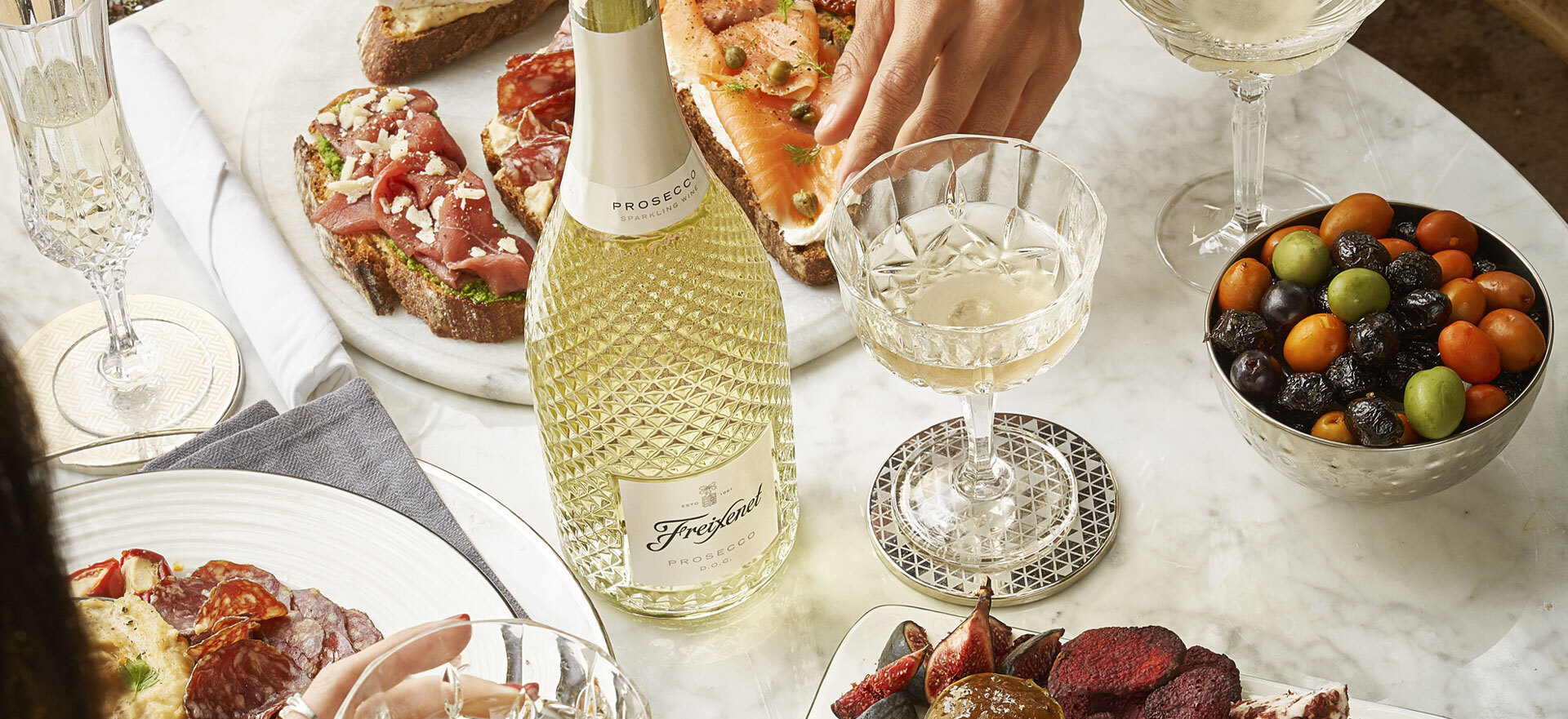 Two women's hands, charcuterie, a faceted sparkling wine bottle and filled coupe glasses.
