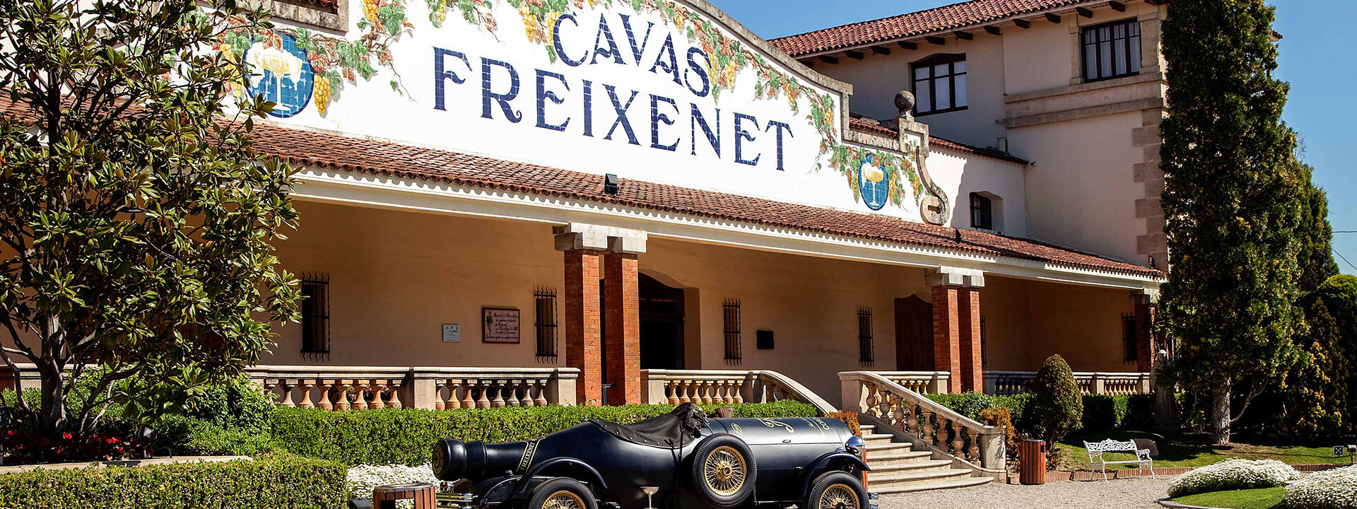 Facade of Freixenet Cava headquarters, a white building with a red roof. In front of it is a car made to look like it is made of a large black wine bottle.